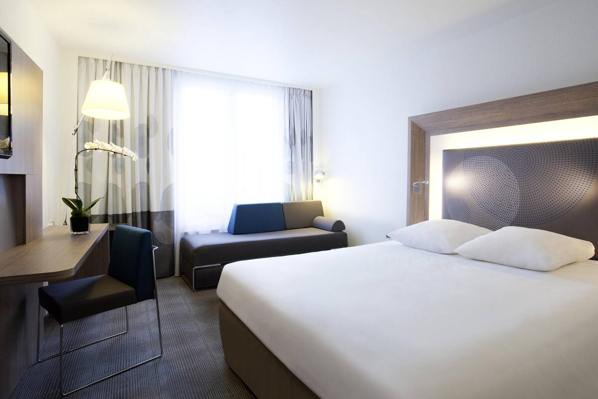 A hotel room with a bed, a desk, a chair, a potted plant, and a window with curtains.