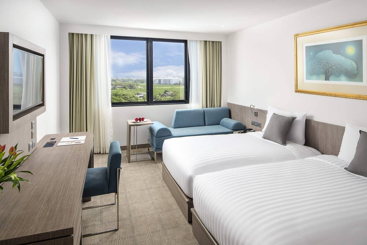 Ett hotellrum med två sängar, ett skrivbord, en stol och ett fönster med utsikt över grönska och byggnader.