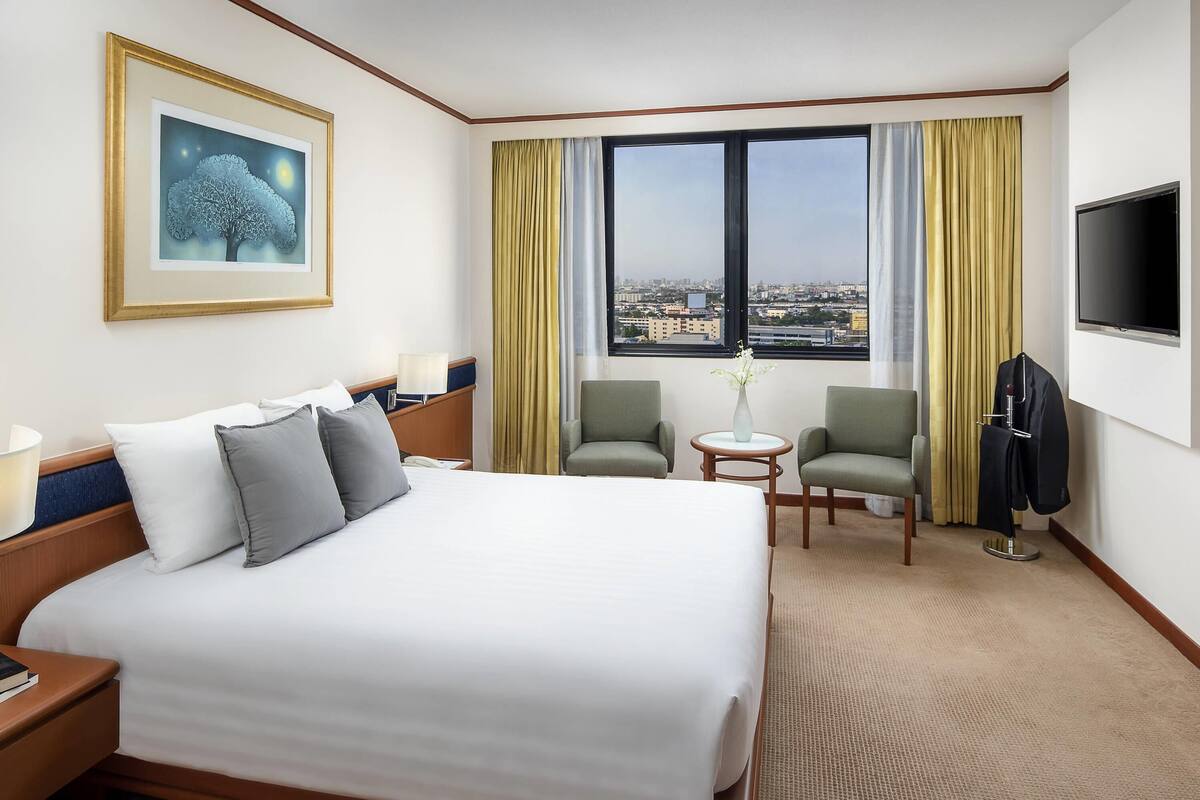 A hotel room with a large bed, two armchairs, a small table, a TV, and a window with a city view.