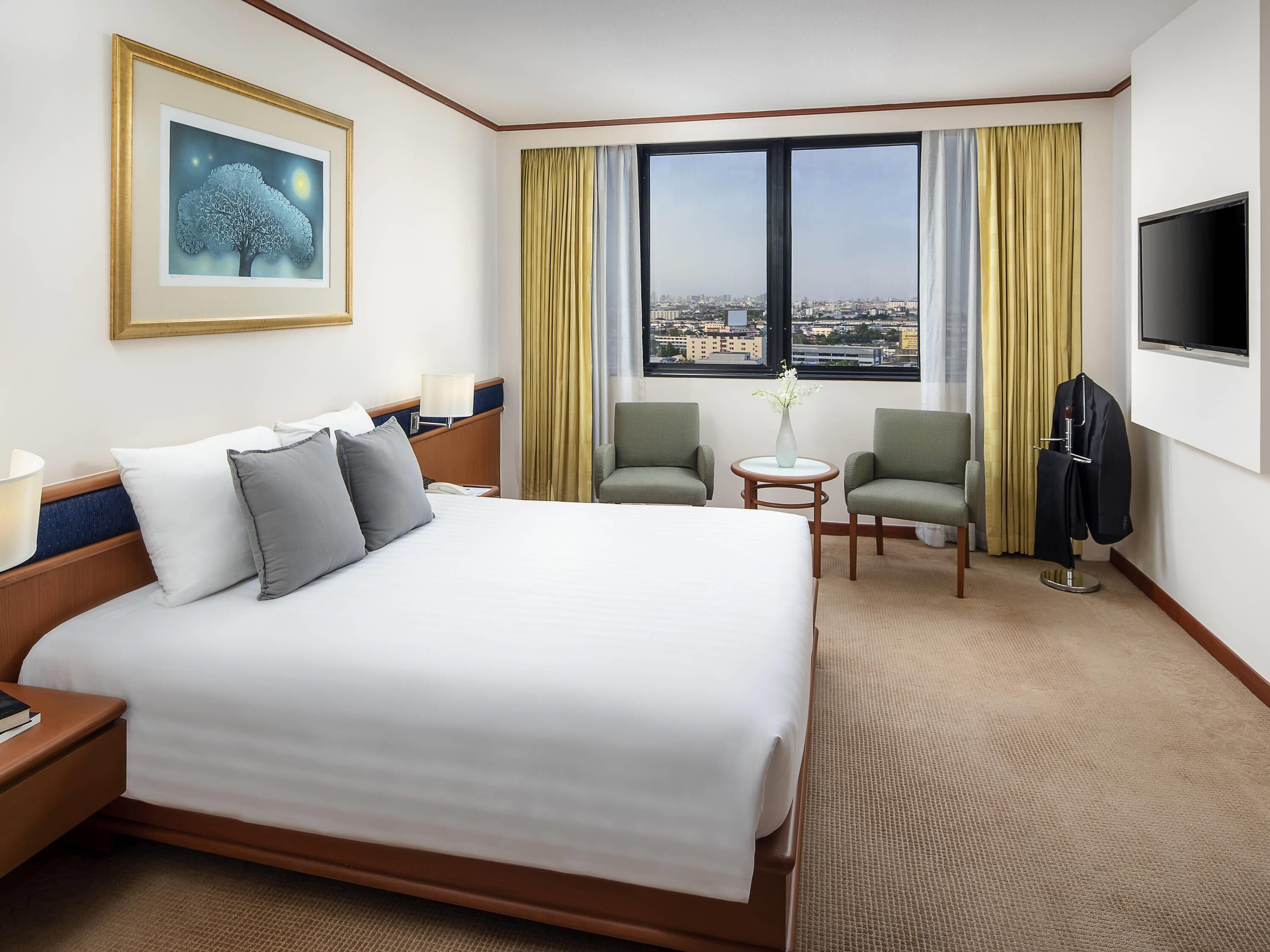 Ein Hotelzimmer mit einem großen Bett, zwei Sesseln, einem kleinen Tisch, einem Fernseher und einem Fenster mit Blick auf die Stadt.
