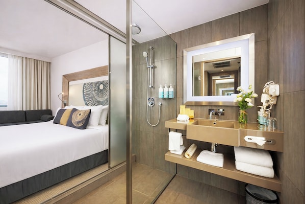 A hotel room with a bed, a shower, a vanity with a mirror, and a window with curtains.