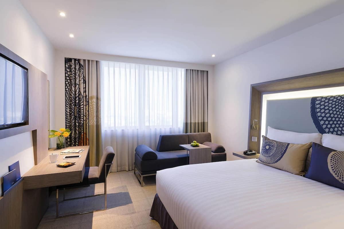 A hotel room with a bed, a desk with a chair, a sofa, a TV, and a window with curtains.