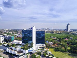A modern hotel with a prominent glass facade, surrounded by greenery and other buildings.