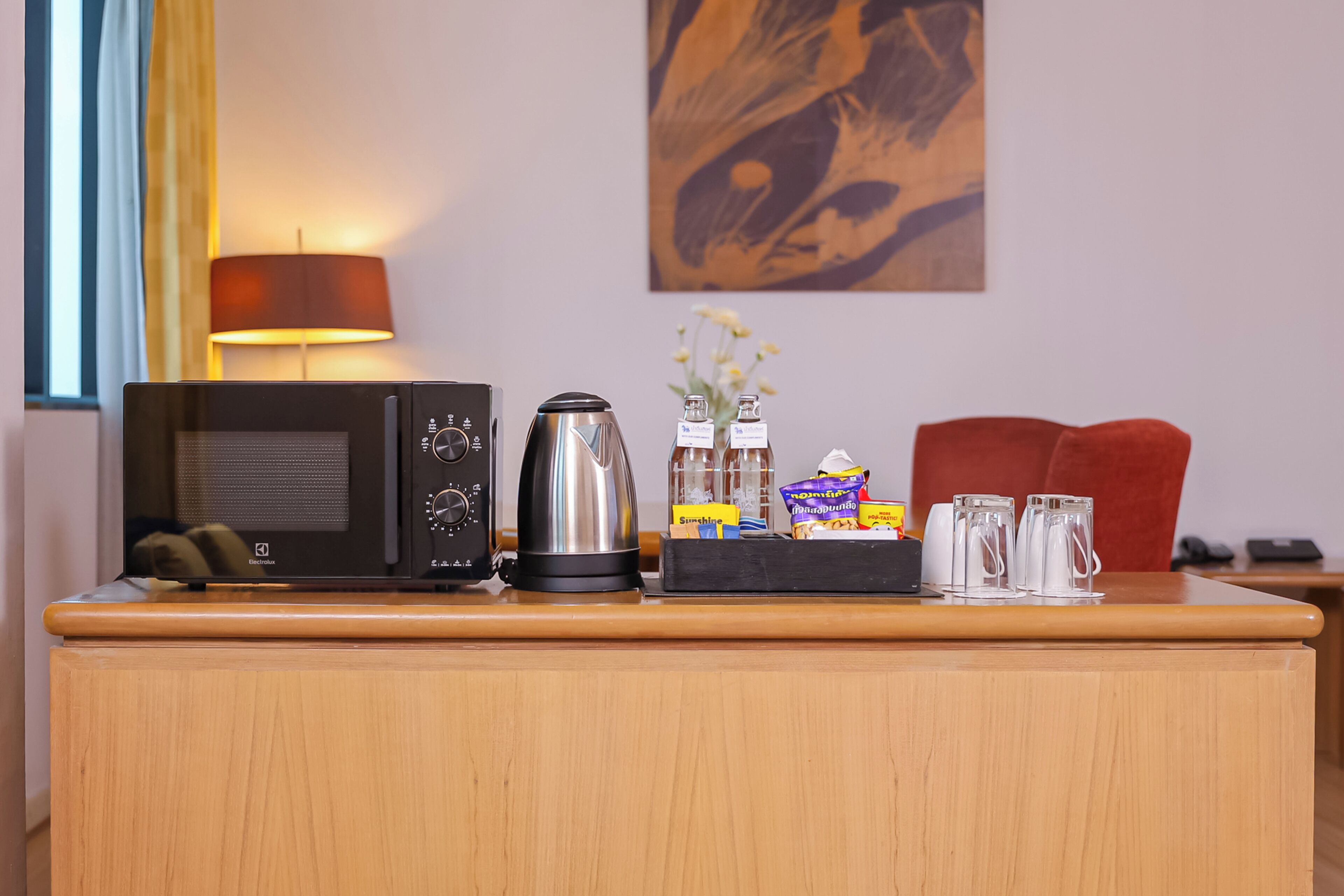 A hotel room desk with a microwave, kettle, and a tray with snacks and glasses.