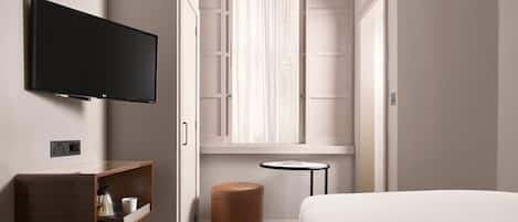 Una habitación de hotel con una cama, un televisor montado en la pared, un pequeño estante de madera con una tetera, un taburete redondo y una ventana con cortinas.