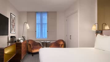 A hotel room with a bed, two armchairs, a small table, a desk with a lamp, a TV, and a window with curtains.