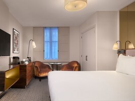 Ein Hotelzimmer mit einem Bett, zwei Sesseln, einem kleinen Tisch, einem Schreibtisch mit Lampe, einem Fernseher und einem Fenster mit Vorhängen.