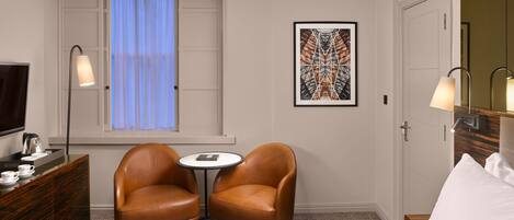 A hotel room with a bed, two leather chairs, a small table, a TV, a desk with a lamp, a framed artwork, and a window with curtains.
