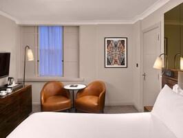 A hotel room with a bed, two leather chairs, a small table, a TV, a desk with a lamp, a framed artwork, and a window with curtains.