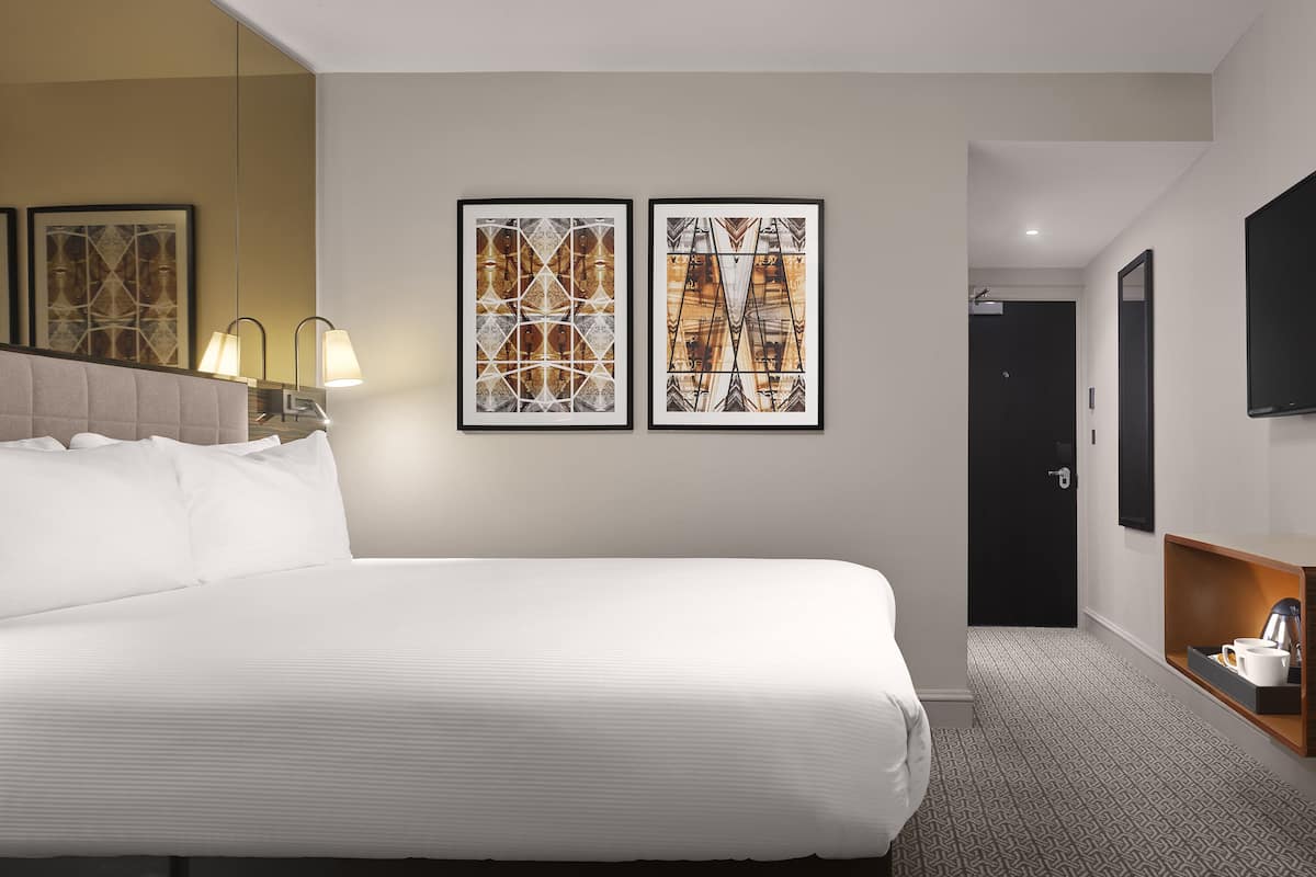 Ett hotellrum med en stor säng, två inramade konstverk på väggen, en sänglampa, en platt-TV och ett litet bord med en kaffebryggare.