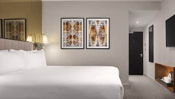 A hotel room with a large bed, two framed artworks on the wall, a bedside lamp, a flat-screen TV, and a small table with a coffee maker.