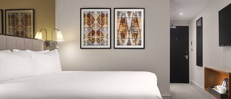 A hotel room with a large bed, two framed artworks on the wall, a bedside lamp, a flat-screen TV, and a small table with a coffee maker.