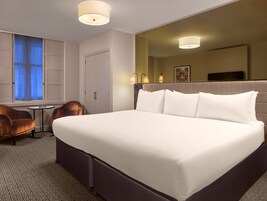Una habitación de hotel con una cama grande, una mesita redonda pequeña, dos sillones, un ventanal con cortinas azules y un televisor montado en la pared.