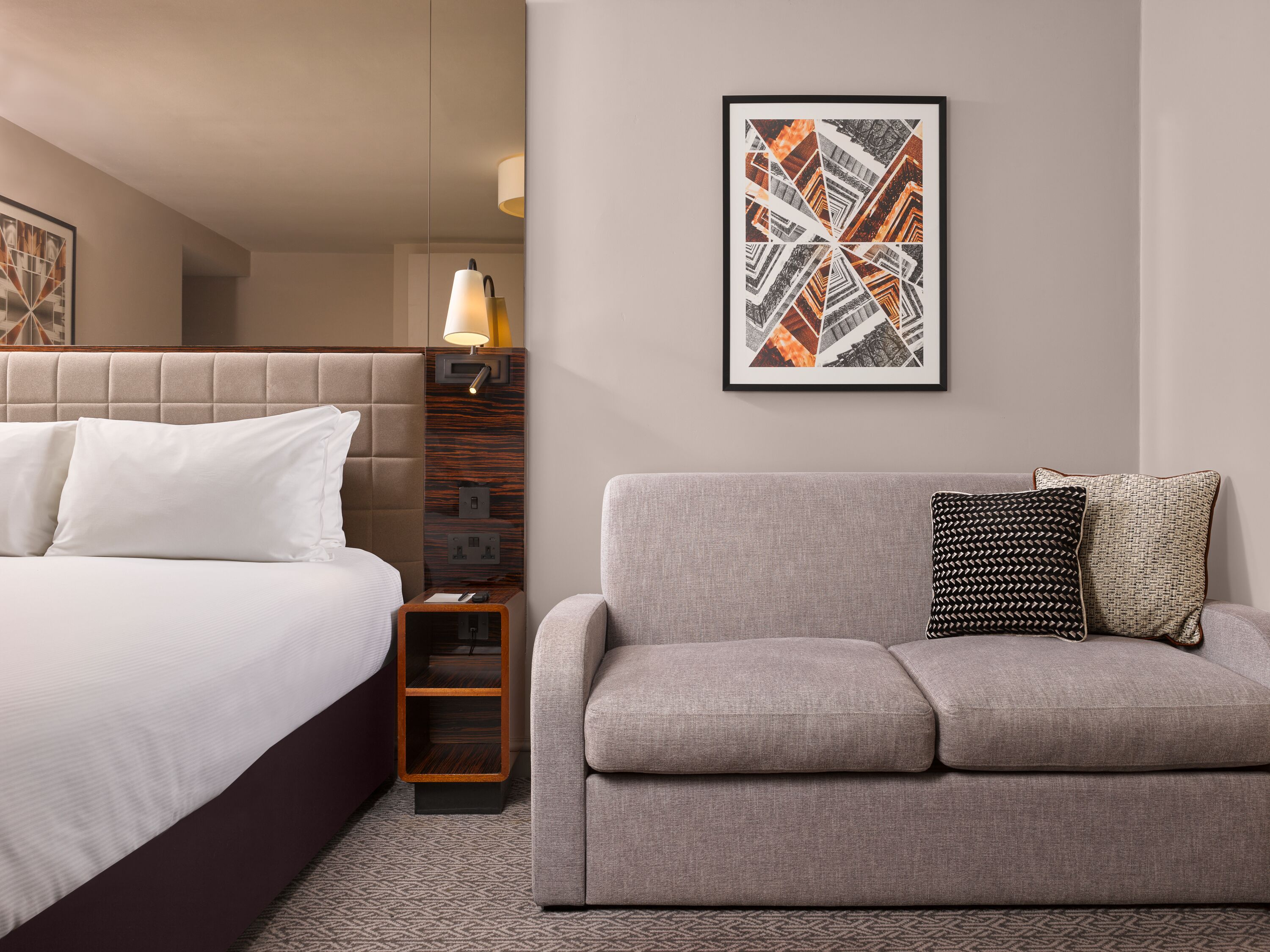 A hotel room with a bed, a sofa, a nightstand, a lamp, and a framed abstract artwork on the wall.