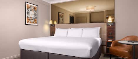 A hotel room with a large bed, a chair, a small table, and a mirror.