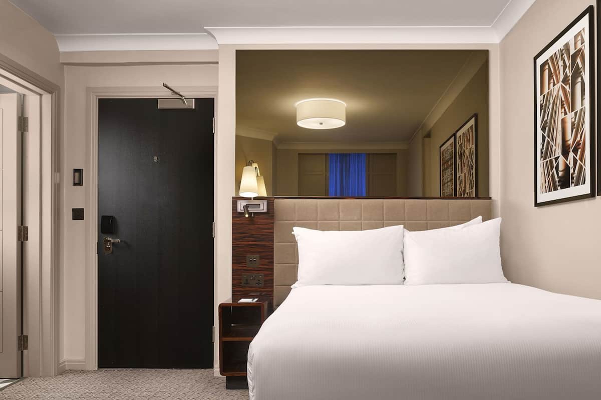 Ett hotellrum med en säng, en dörr, en lampa och en vägg med inramade konstverk.