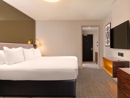 Camera d'albergo con un letto grande, lampade sui comodini, una TV a schermo piatto, una cassettiera in legno e pavimento con moquette.