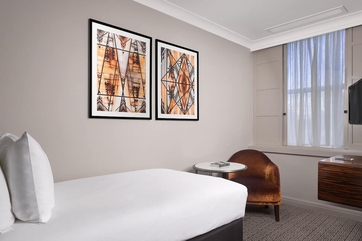 Ett hotellrum med en säng, en stol, ett litet bord och två inramade konstverk på väggen.