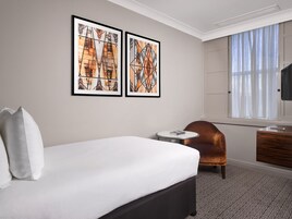 Ein Hotelzimmer mit einem Bett, einem Sessel, einem kleinen Tisch und zwei gerahmten Kunstwerken an der Wand.