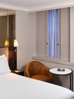 Ein Hotelzimmer mit einem Bett, einem braunen Ledersessel, einem kleinen runden Tisch, einem Nachttisch, einer Lampe, einem Spiegel und einem Fenster mit Vorhängen.