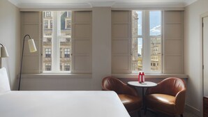 A hotel room with a bed, two leather chairs, a small round table, a floor lamp, and large windows with a city view.