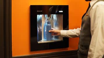 Eine Person bedient ein Wasserautomaten der Marke ELKAY vor einer orangefarbenen Wand.