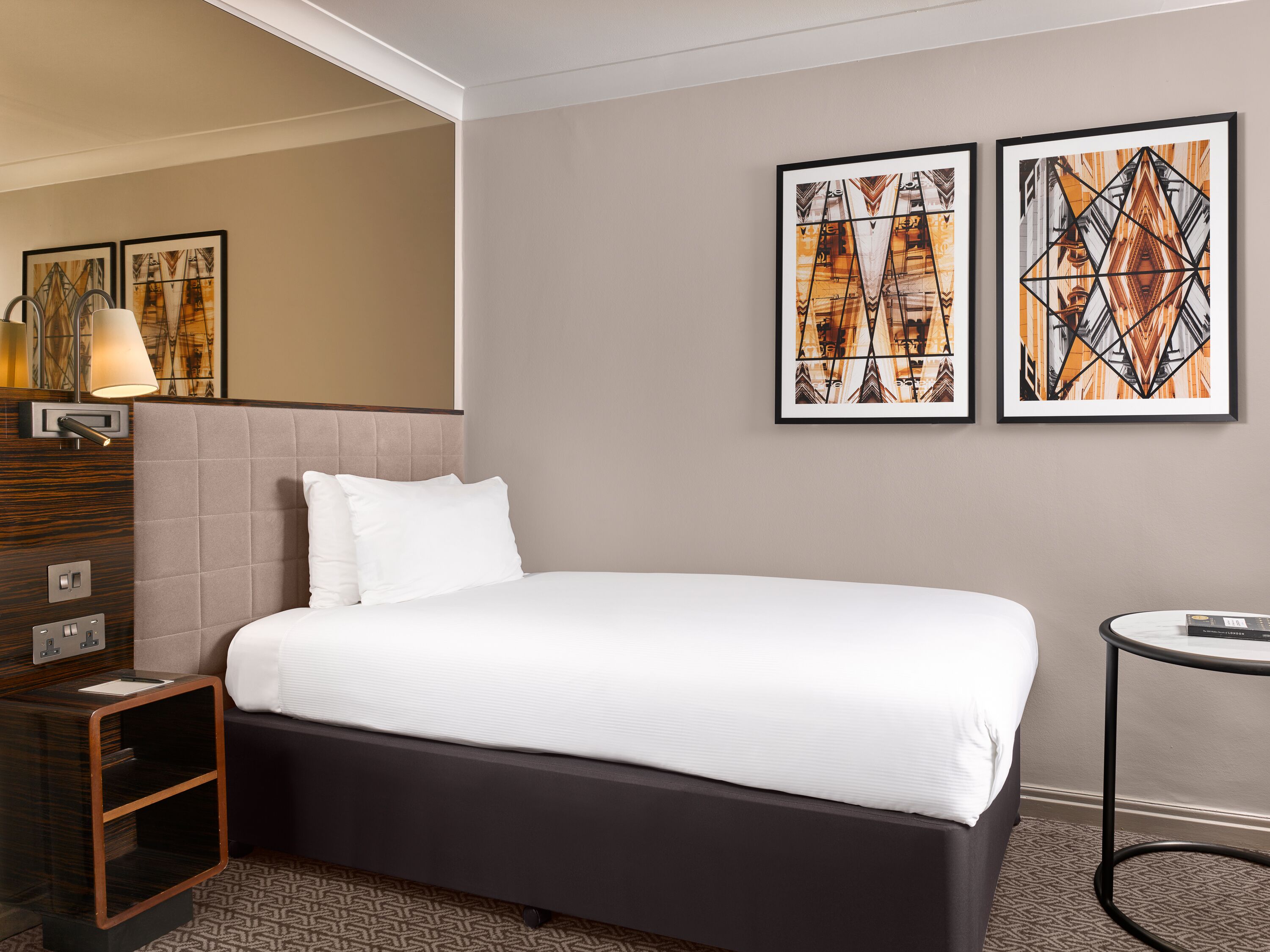 A hotel room with a large bed, a bedside table, a lamp, and two framed artworks on the wall.