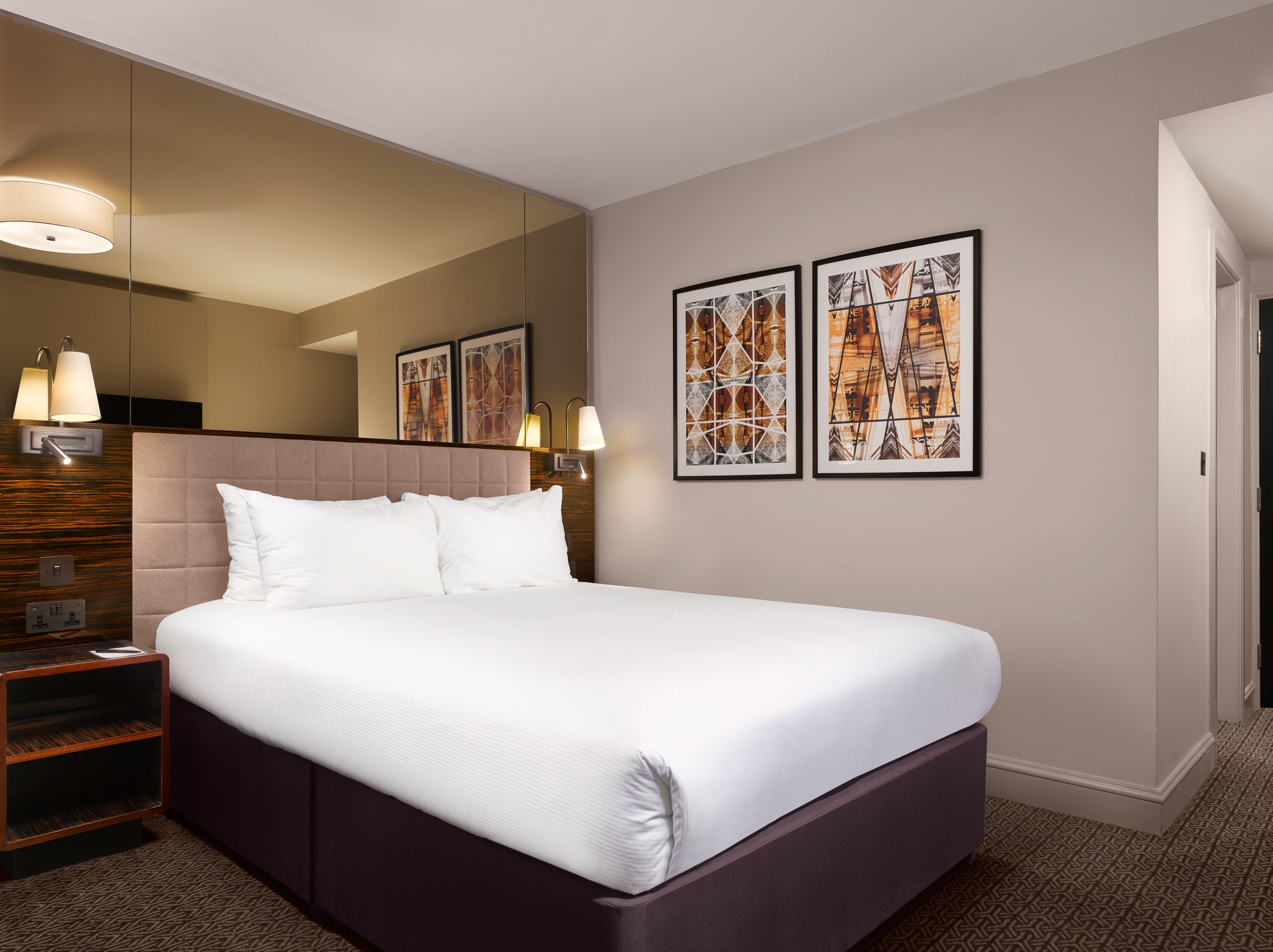Habitación de hotel con una cama grande, una mesita de noche, un espejo y dos cuadros en la pared.