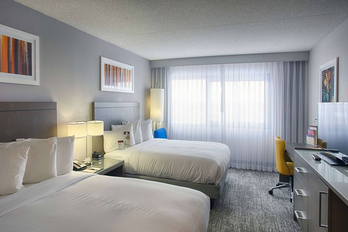 Une chambre d’hôtel avec deux lits, un bureau, une chaise et une grande fenêtre avec des rideaux légers.