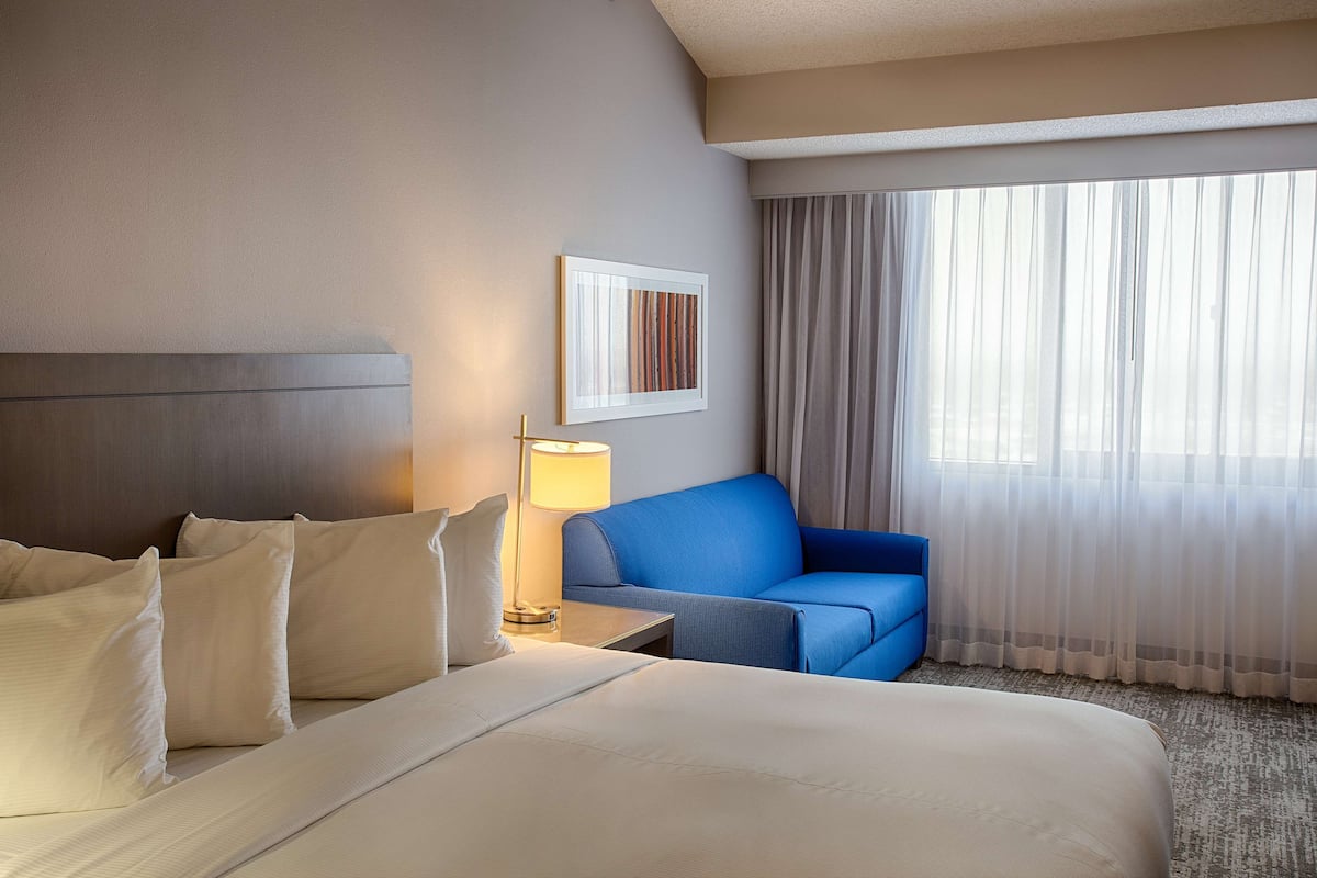 A hotel room with a large bed, a blue armchair, a bedside table with a lamp, and a large window with curtains.