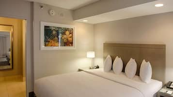 A hotel room with a large bed, a headboard, a bedside table with a lamp, a framed artwork on the wall, and a door leading to another room.