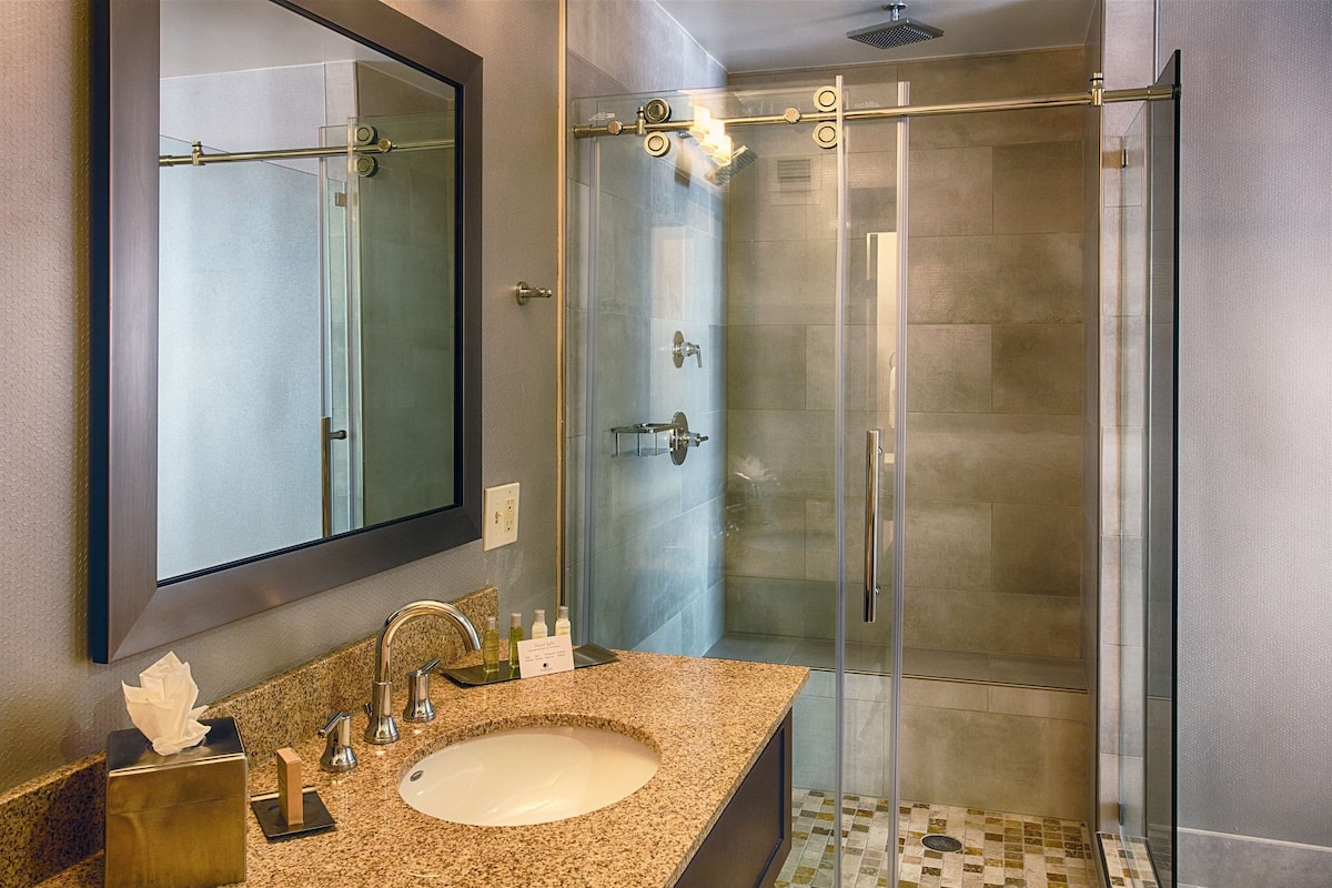 A modern bathroom with a granite countertop, a large mirror, a glass-enclosed shower, and a sink with a towel dispenser.