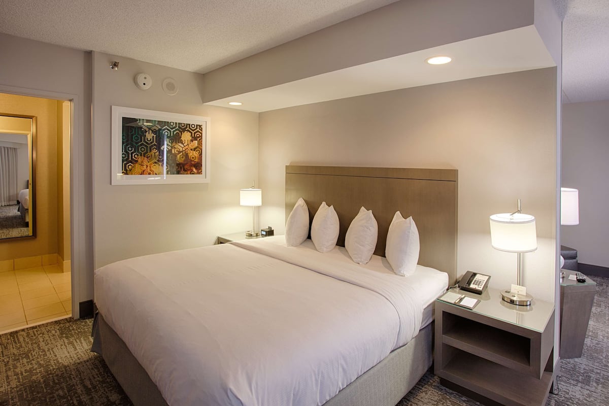 A hotel room with a large bed, two bedside tables with lamps, a headboard, a framed artwork on the wall, and a carpeted floor.