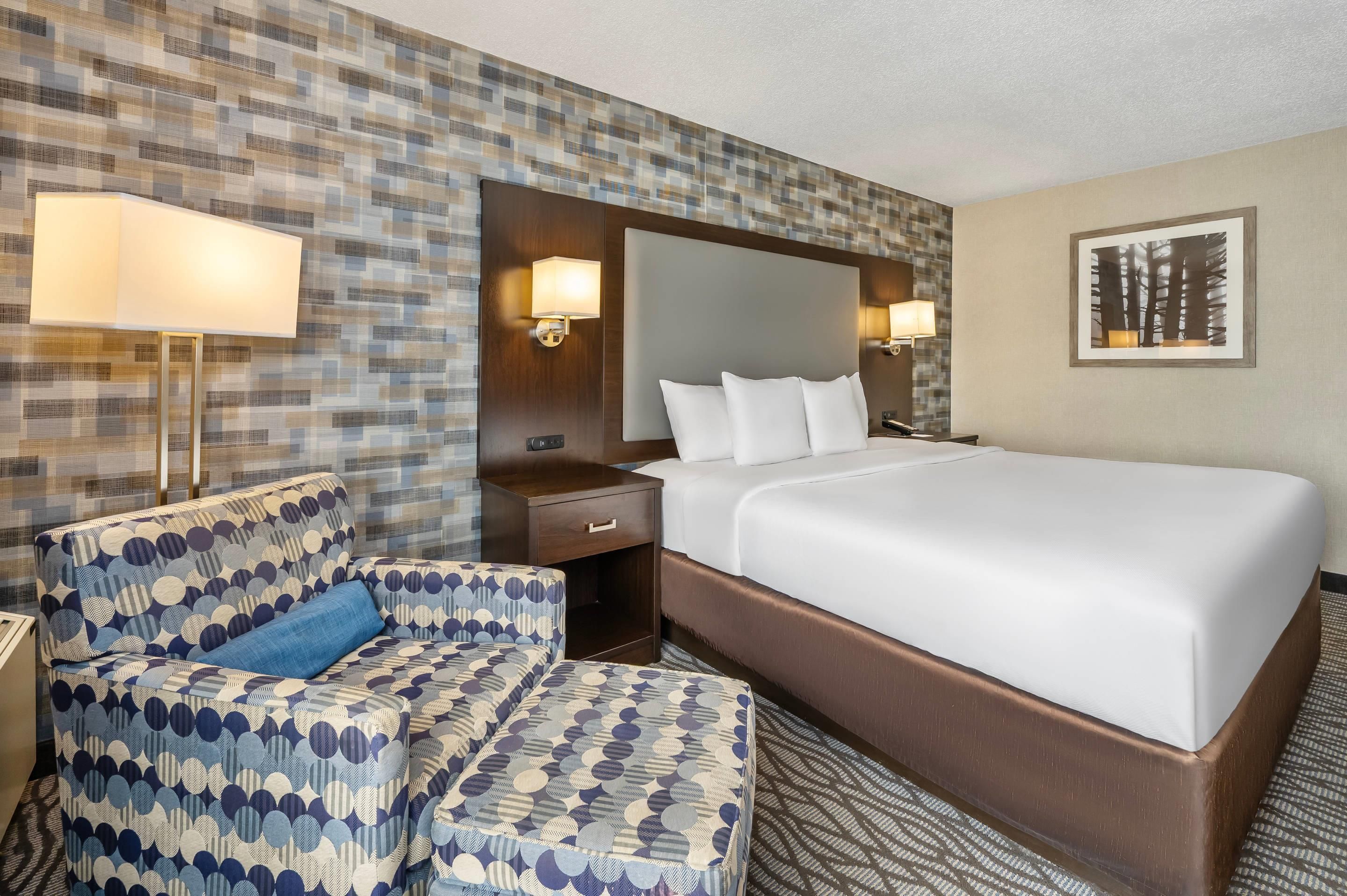 A hotel room with a large bed, a patterned armchair, a nightstand, and two wall-mounted lamps.