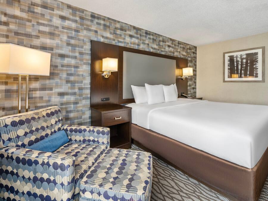 Una habitación de hotel con una cama grande, un sillón estampado, una mesita de noche y dos lámparas fijadas a la pared.