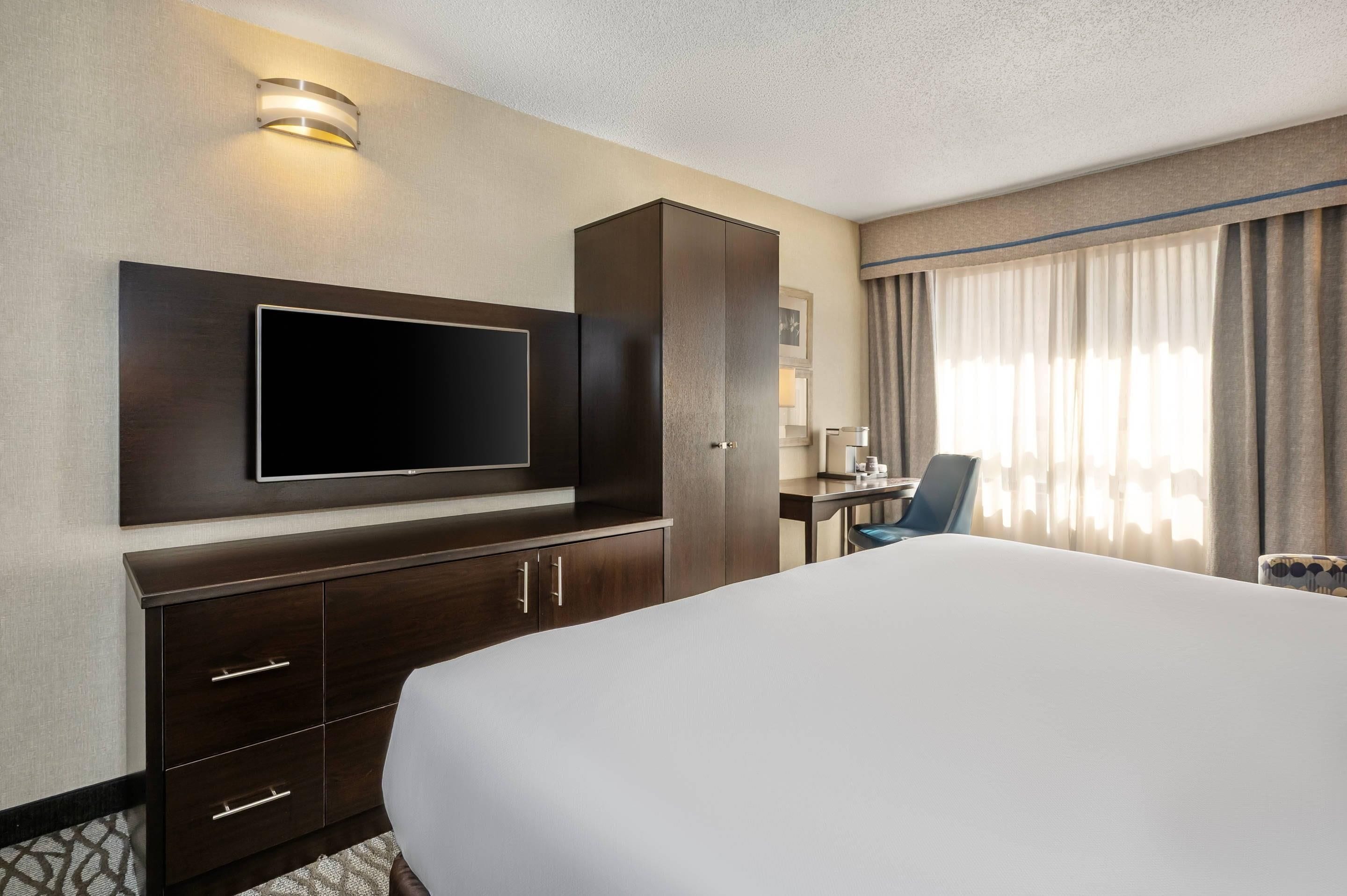A hotel room with a large bed, a flat-screen TV mounted on the wall, a desk with a chair, and a window with curtains.