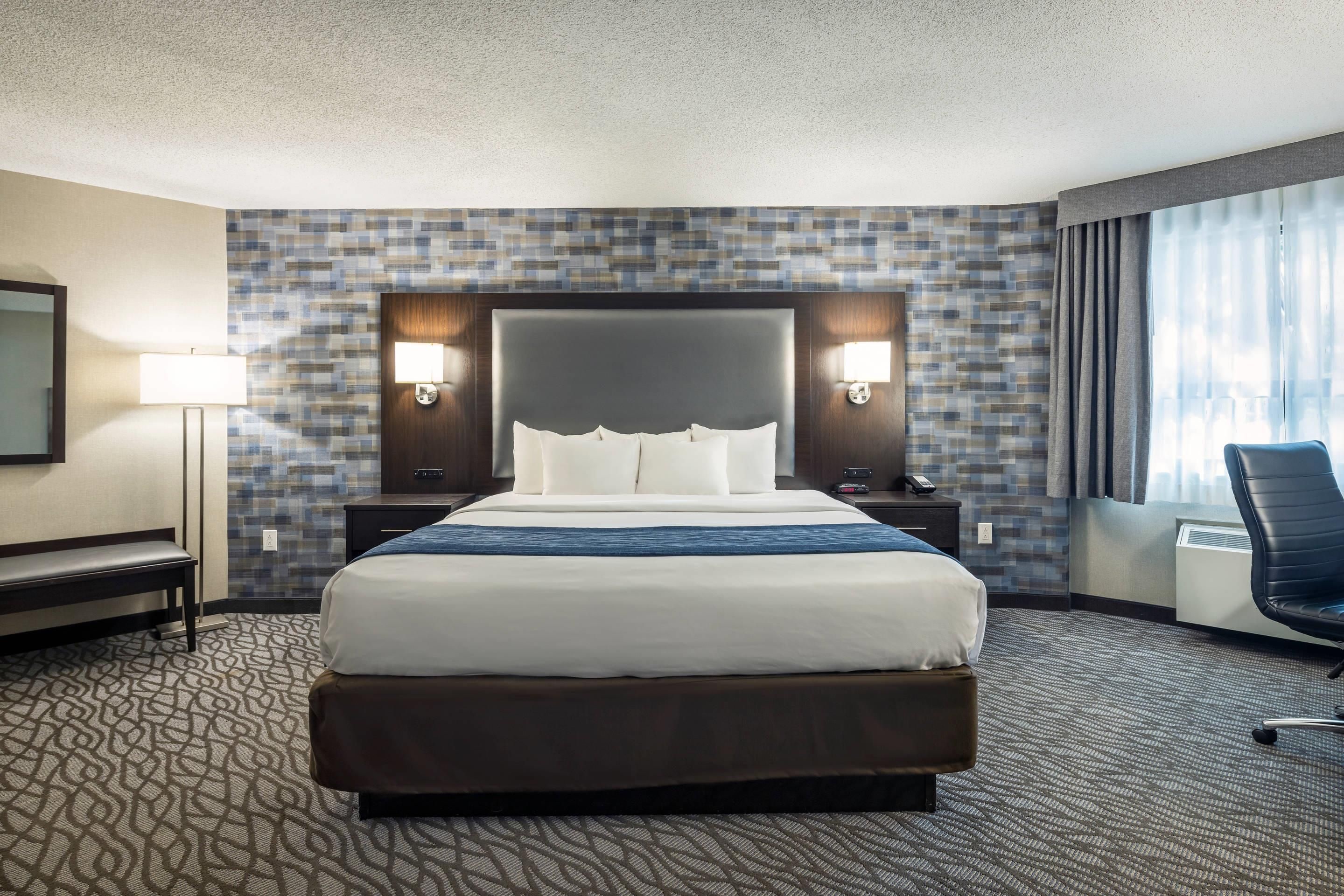 Ein Hotelzimmer mit einem großen Bett, zwei Nachttischen, einem Schreibtisch, einem Stuhl, einem Spiegel und einem Fenster mit Vorhängen.