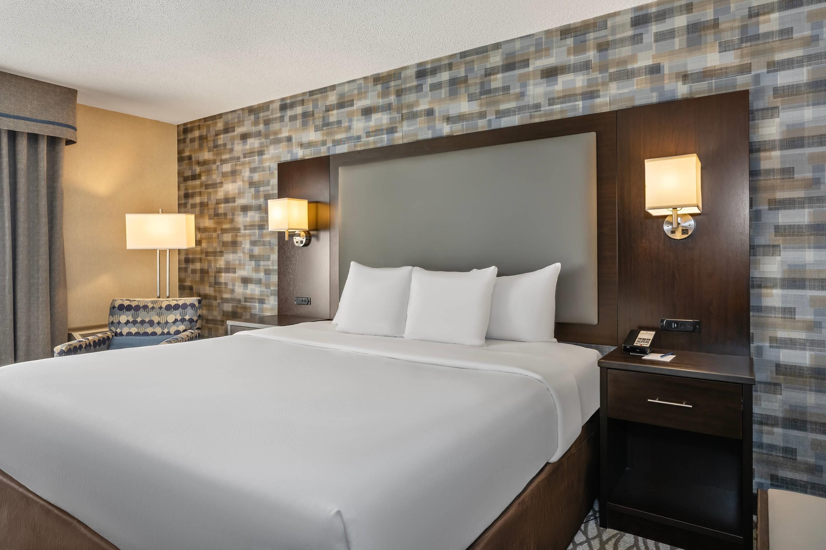 A hotel room with a large bed, two bedside lamps, a nightstand, a patterned wall, and a window with curtains.
