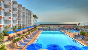 A pool area with lounge chairs and umbrellas, a clear view of the ocean, and a building with balconies.