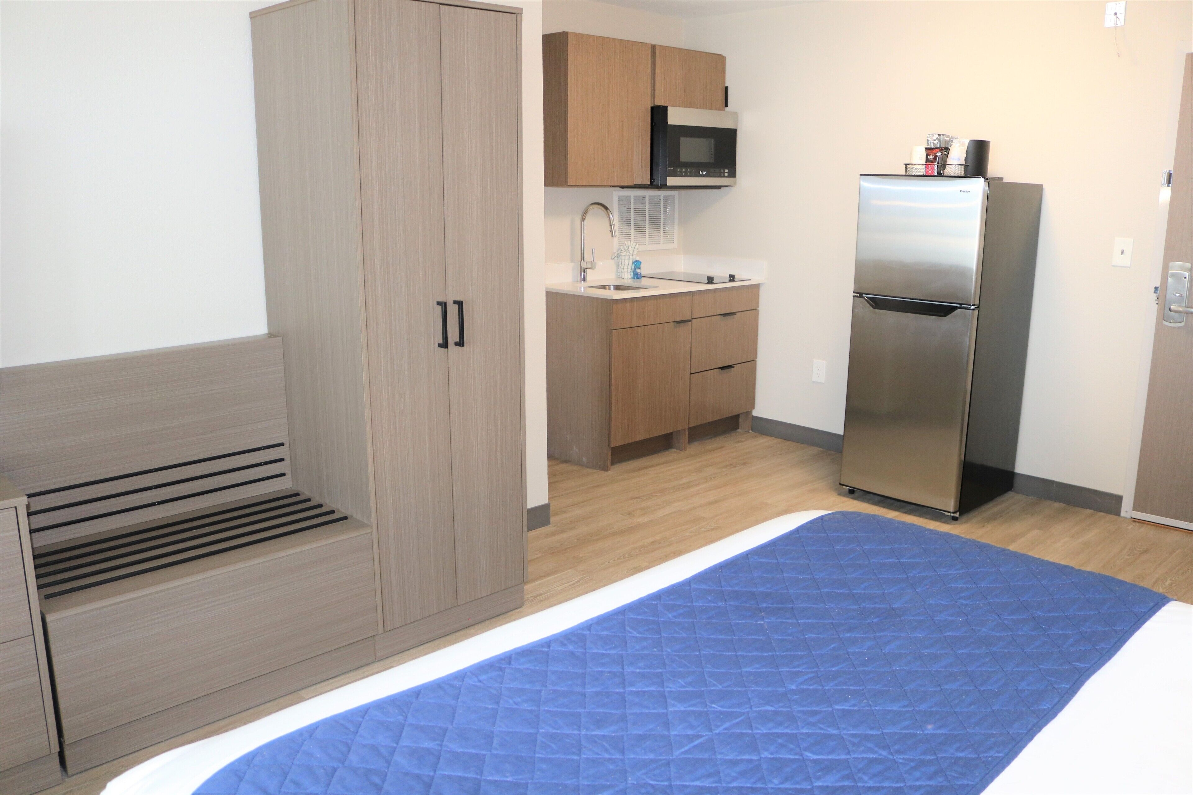 Una habitación compacta con cama, armario empotrado, kitchenette con microondas y refrigerador, y colcha acolchada azul.