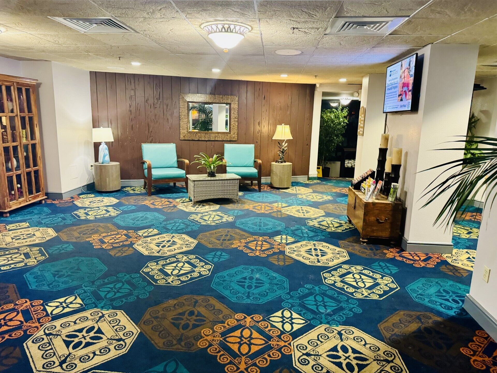 A lobby with a patterned carpet, wooden walls, two blue armchairs, a small table with a plant, and a television screen.