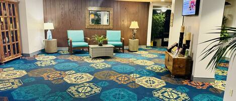 A lobby with a patterned carpet, wooden walls, two blue armchairs, a small table with a plant, and a television screen.