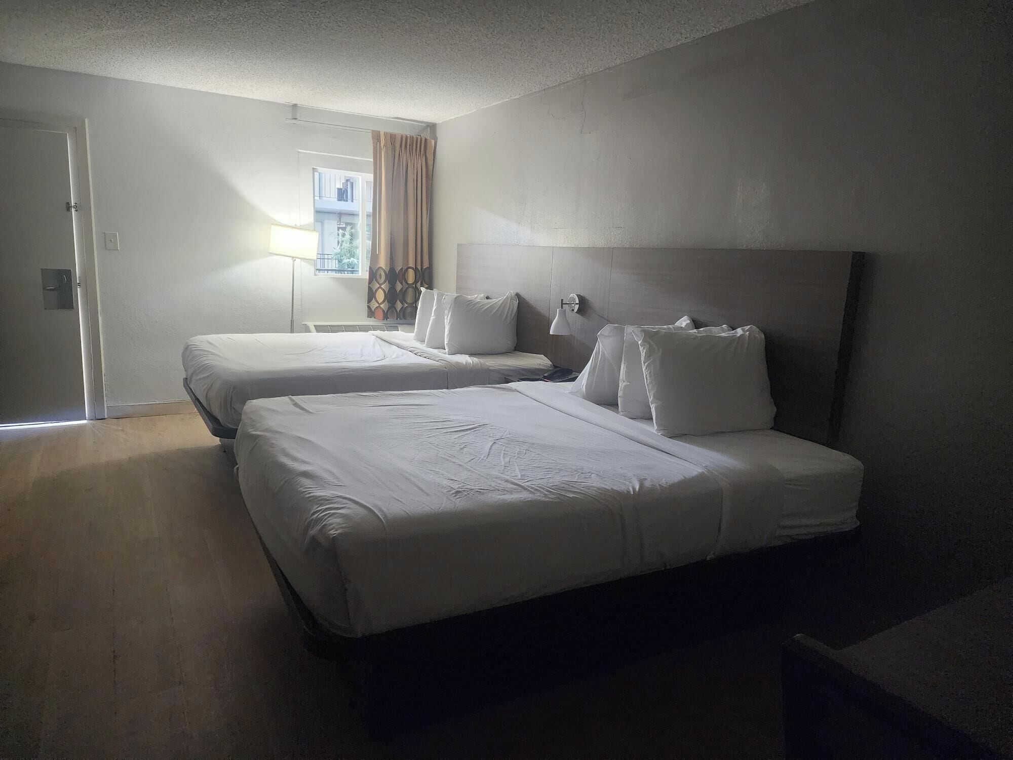 Habitació d'hotel amb dos llits individuals, una finestra gran amb cortines, una làmpada de nit i una porta.