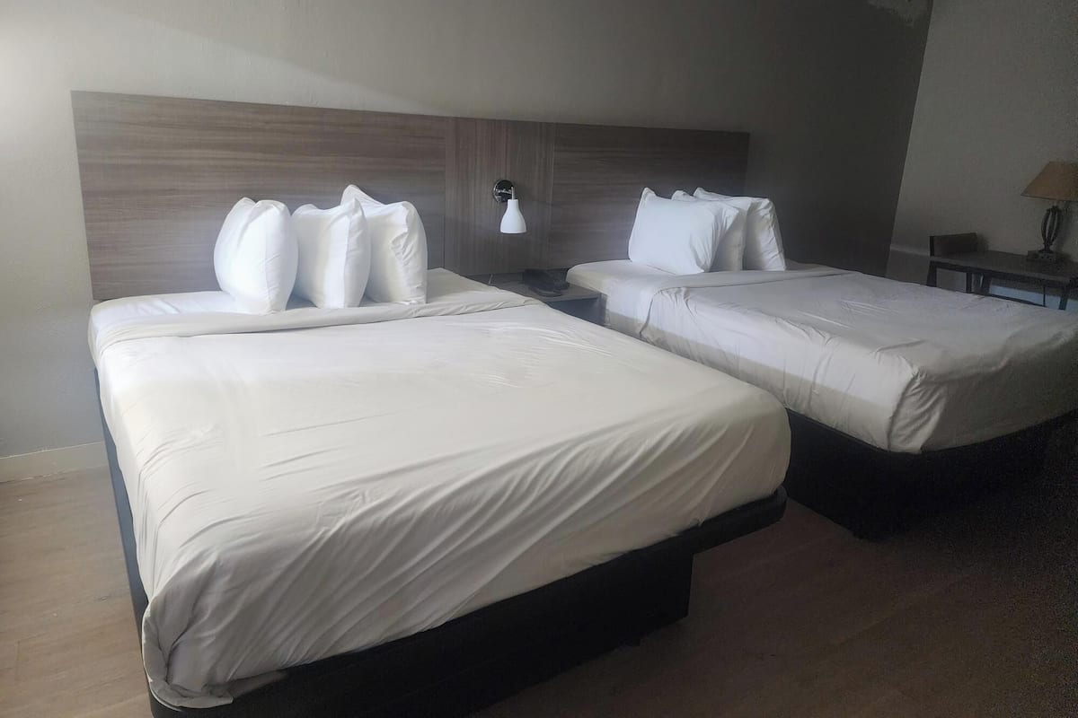 A hotel room with two beds, a wooden headboard, a nightstand with a lamp, and a small table with a lamp.
