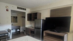 Een hotelkamer met een bed, een bureau met stoel, een kitchenette met koelkast en een televisie op een standaard.