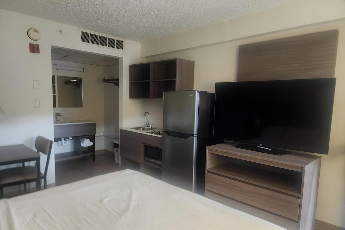 A hotel room with a bed, a desk with a chair, a kitchenette with a refrigerator and a television on a stand.