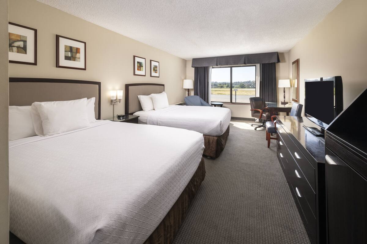 A hotel room with two beds, a desk, a TV, and a view of the outdoors.