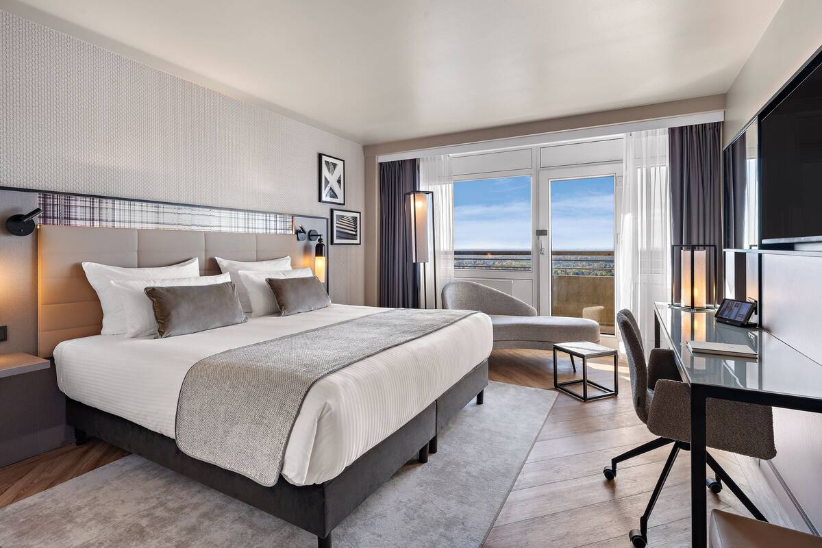 Une chambre d’hôtel moderne dotée d’un grand lit, d’un bureau, d’une chaise et d’un balcon offrant une vue sur la ville.