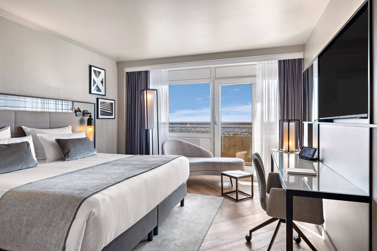 Een moderne hotelkamer met een groot bed, een bureau met een computer, een stoel en uitzicht op de stad door grote ramen.