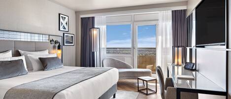 Una habitación de hotel moderna con una cama grande, un escritorio con computadora, una silla y vistas a la ciudad a través de amplias ventanas.